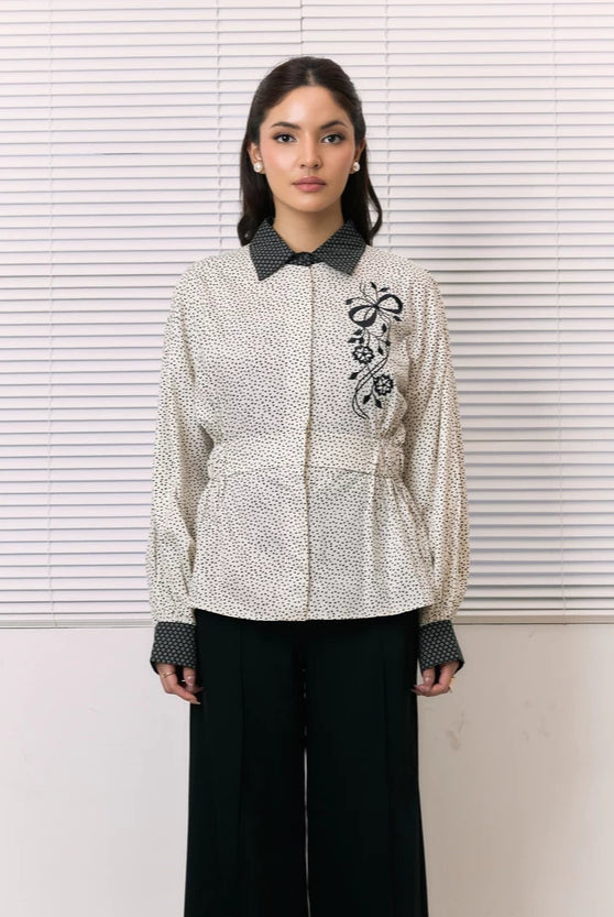 A woman stands in front of white window blinds wearing a cream-colored textured shirt by Studio Tui with black floral embroidery on the chest, a black patterned collar and cuffs, and black wide-leg pants.