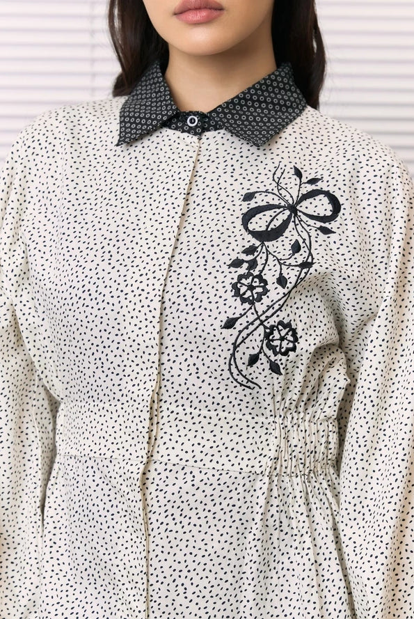 A close-up of a woman wearing a cream-colored long-sleeve shirt by Studio Tui with a black micro-dot pattern, a contrasting black patterned collar, and a black floral embroidery on the chest.