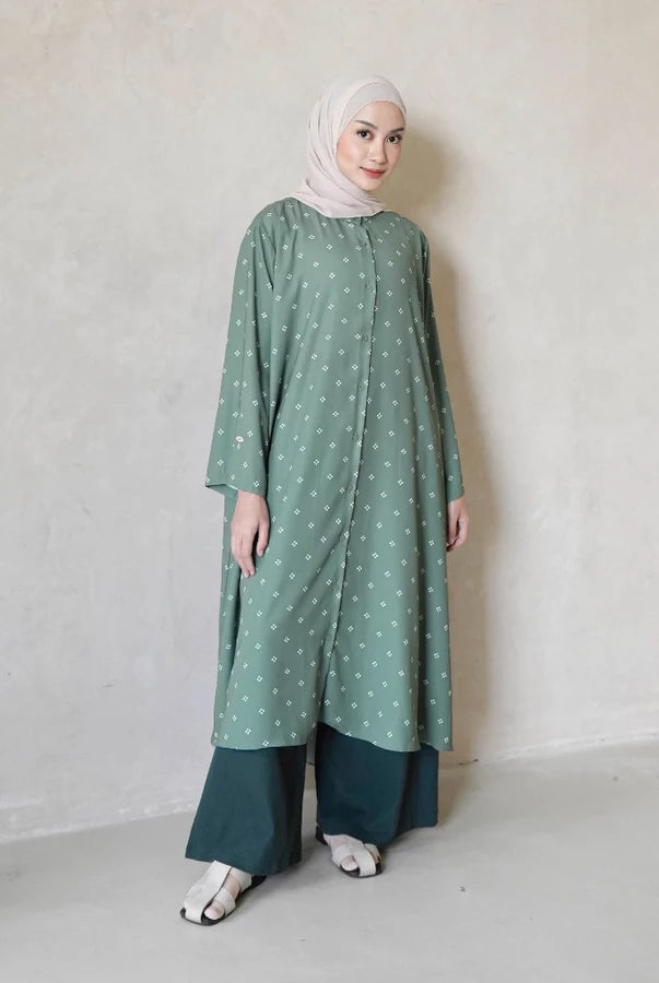 Woman wearing a green charcoal tunic with white patterns by Mistyvori and a hijab against a plain background
Seorang wanita yang mengenakan tunik berwarna hijau arang dengan motif putih dari Mistyvori dan hijab, dengan latar belakang polos