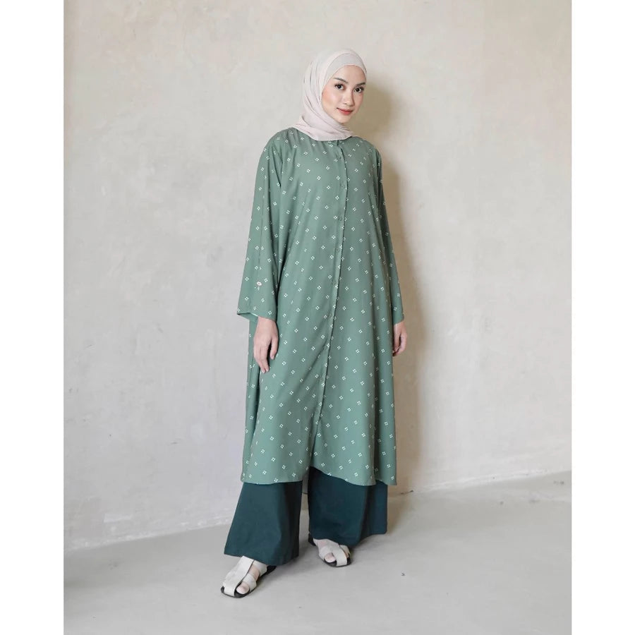 Woman wearing a green charcoal tunic with white patterns by Mistyvori and a hijab against a plain background
Seorang wanita yang mengenakan tunik berwarna hijau arang dengan motif putih dari Mistyvori dan hijab, dengan latar belakang polos