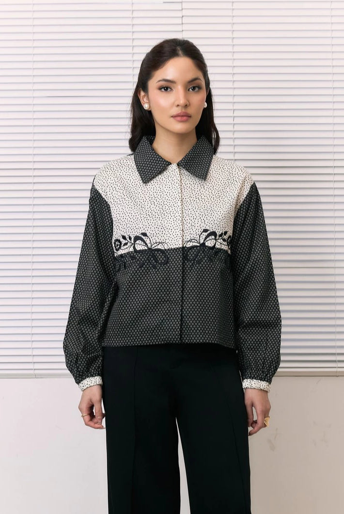 A woman stands in front of white window blinds wearing a cropped long-sleeve shirt by Studio Tui with a black-and-white patterned body, contrasting solid black collar and sleeves, and black pants.