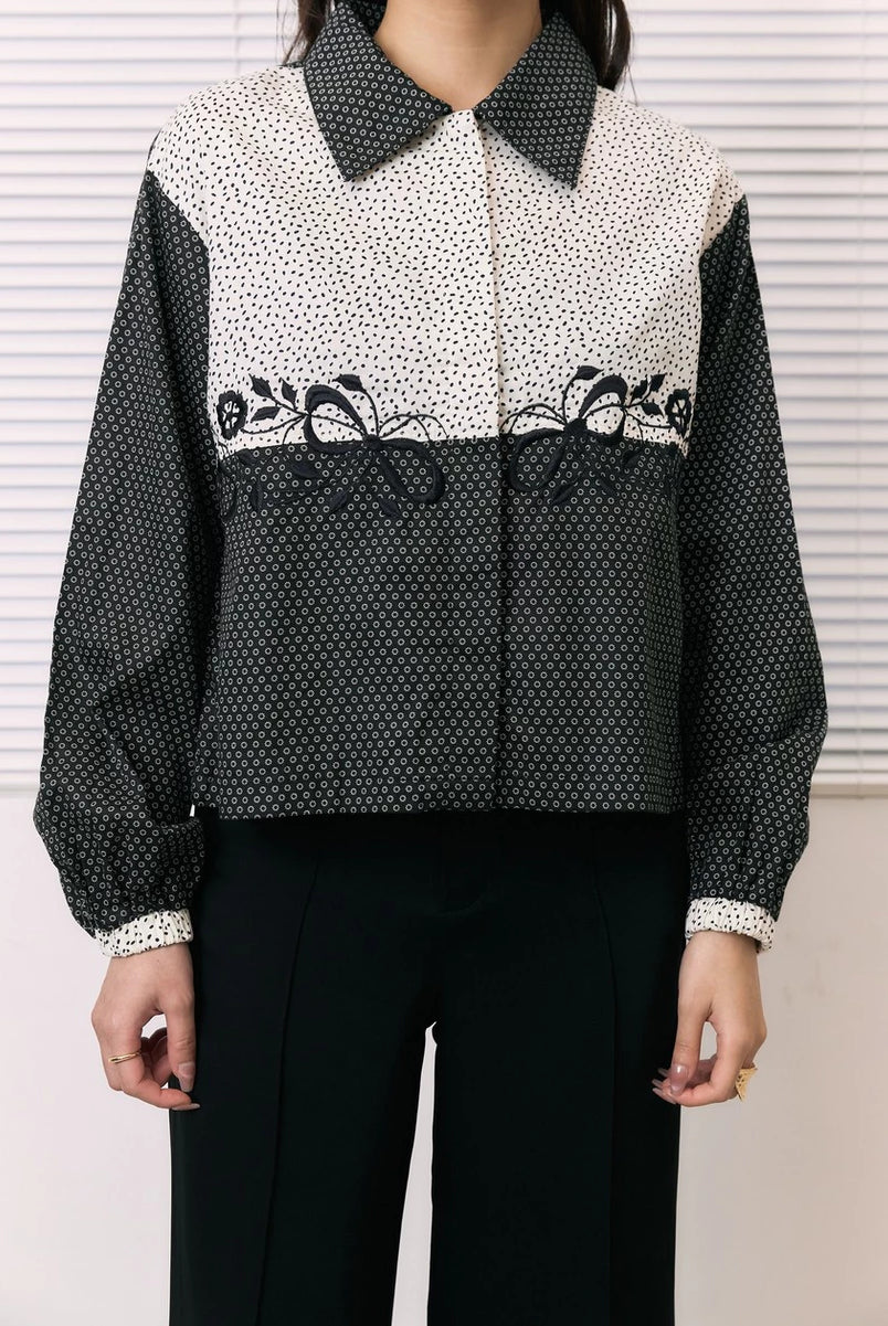 A woman wearing a long-sleeve shirt by Studio Tui with a color-blocked design: a cream and black micro-dot pattern on the upper chest and shoulders, solid black sleeves, and a black-and-white geometric pattern across the lower half. She is also wearing black pants.