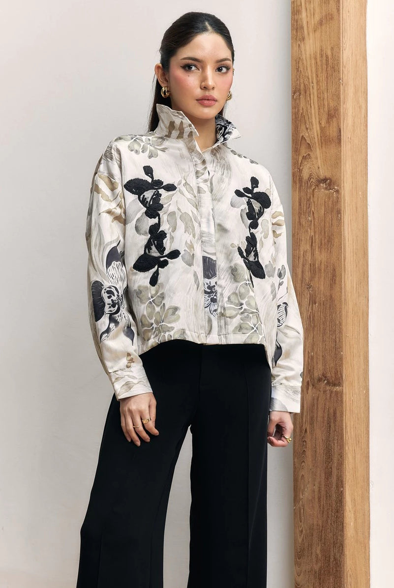 A woman modeling a cropped, long-sleeve high-neck shirt by Studio Tui with a black, gray, and white floral pattern on a cream background, paired with black wide-leg pants, standing next to a wooden pillar.