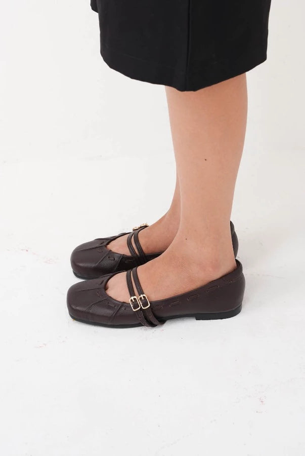 A person wearing dark brown leather Mary Jane flatshoes by Cekhas with double straps and small gold buckle details, paired with a black skirt, shown from the knees down on a white studio background.