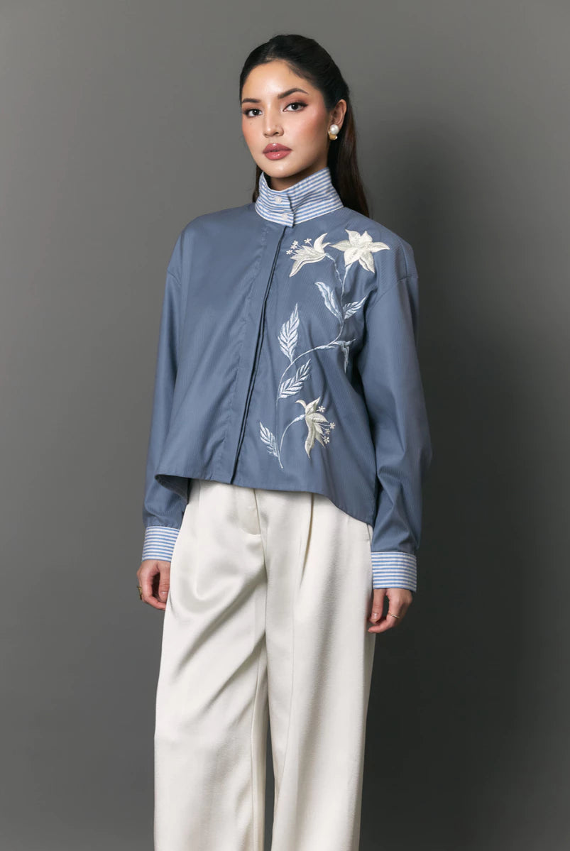A model wearing a dusty blue cropped shirt by Studio Tui with white floral embroidery on the chest, paired with cream wide-leg trousers, against a solid gray studio background.