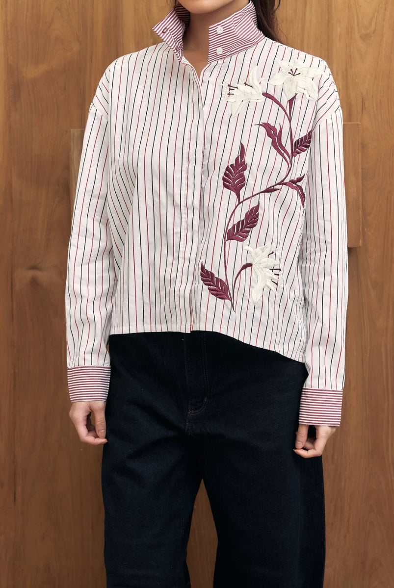 Person wearing a white shirt with red stripes and floral embroidery by Studio Tui against a wooden background