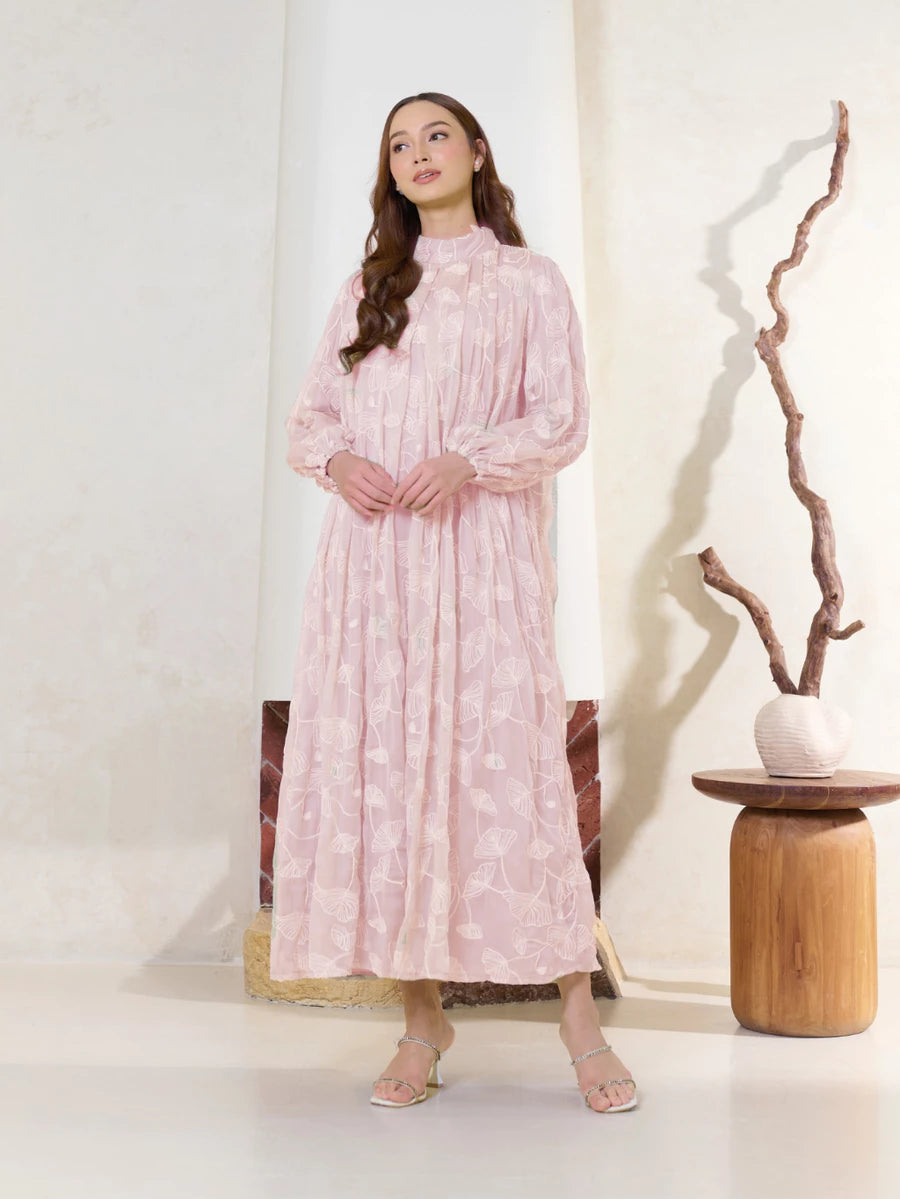 Woman wearing a pink floral dress by Kasa Heritage standing in a minimalistic room with a white wall and wooden table.