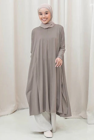 Woman wearing a long, light gray tunic by Mistyvori with a matching hijab against a white curtain background.