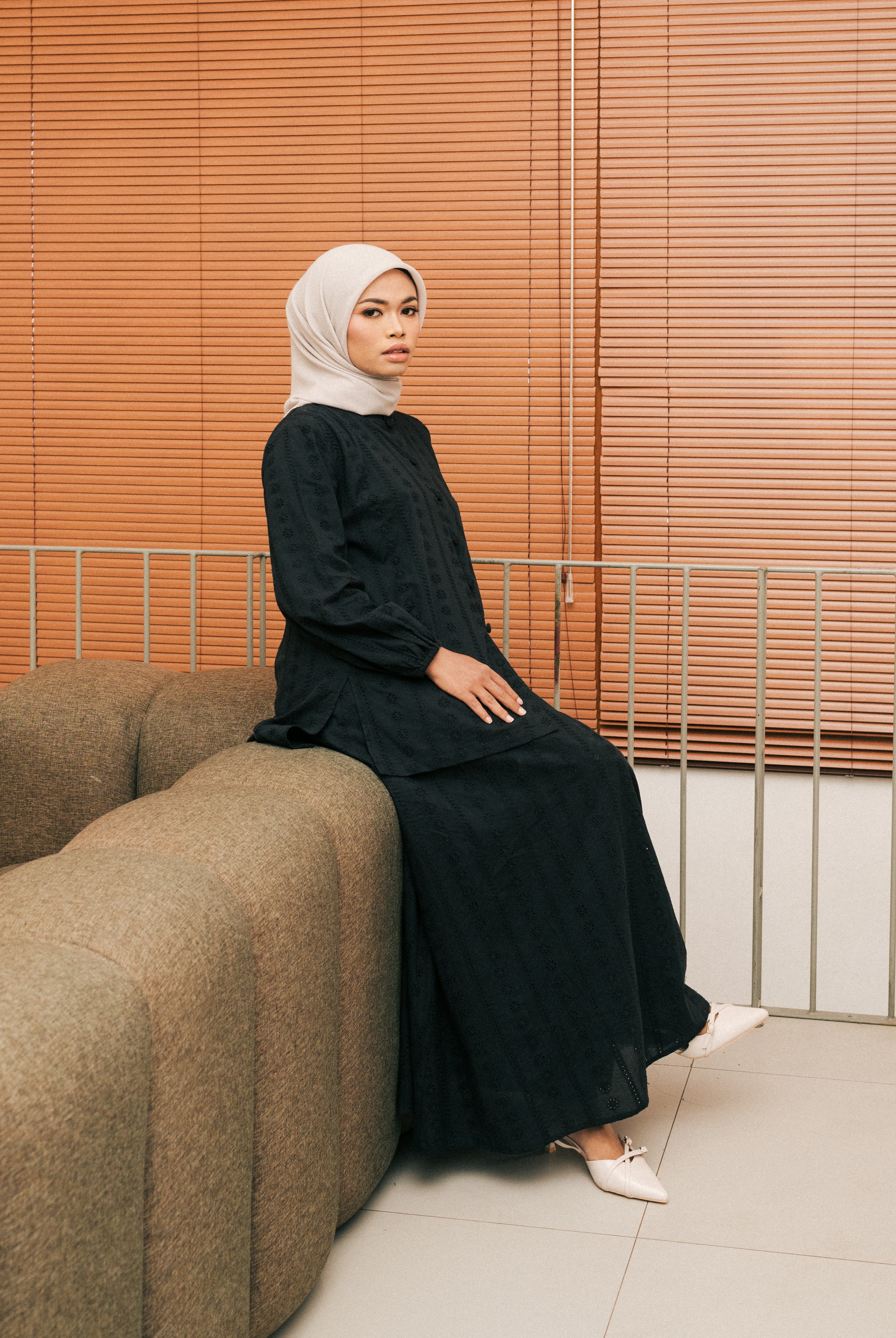 Woman in a black tunic from Atala Scarves and hijab sitting on a brown couch with wooden blinds in the background