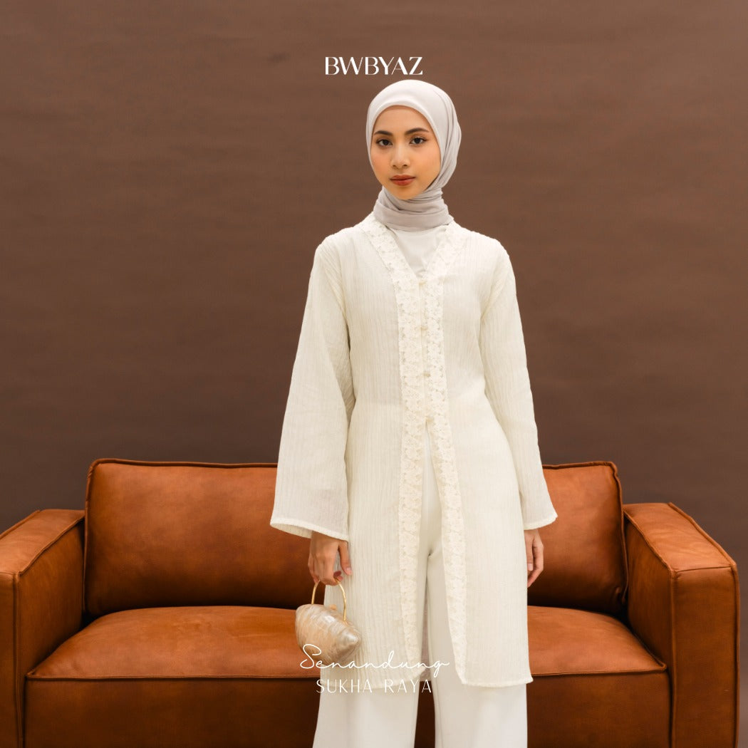 A woman wearing a white long-sleeve floral outer from BWBYAZ with a hijab, standing in front of a brown background.