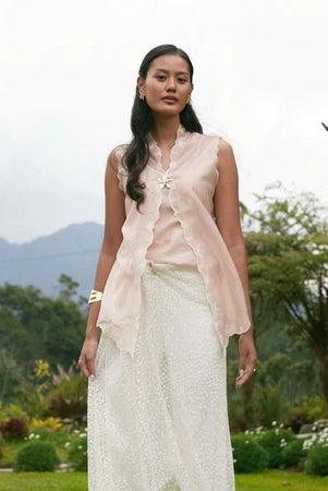 Woman in a light pink sleeveless kebaya by Kartinis Label and white skirt standing outdoors with greenery and mountains in the background
