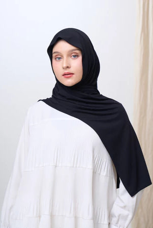 Woman wearing a black pashmina hijab and white top against a plain background