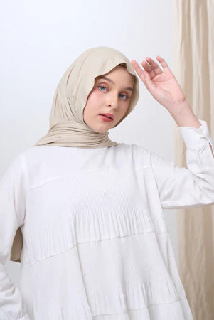Woman wearing a beige pashmina hijab and white outfit against a neutral background