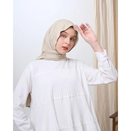 Woman wearing a beige pashmina hijab and white outfit against a neutral background