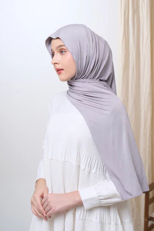 Woman wearing a light gray pashmina hijab and white dress against a plain background