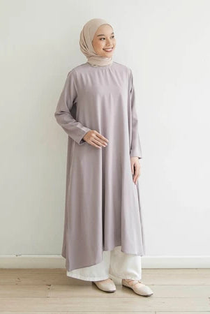 Seorang wanita memakai tunik berlengan panjang berwarna lavender, hijab, dan celana panjang.