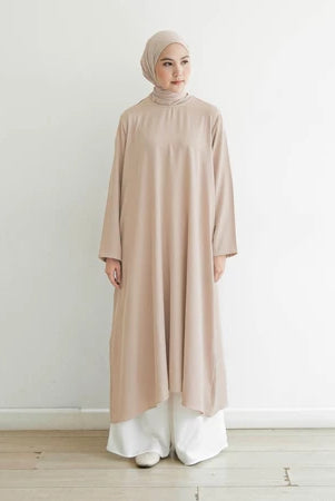 Seorang wanita memakai tunik berlengan panjang berwarna beige muda, hijab, dan celana panjang.
