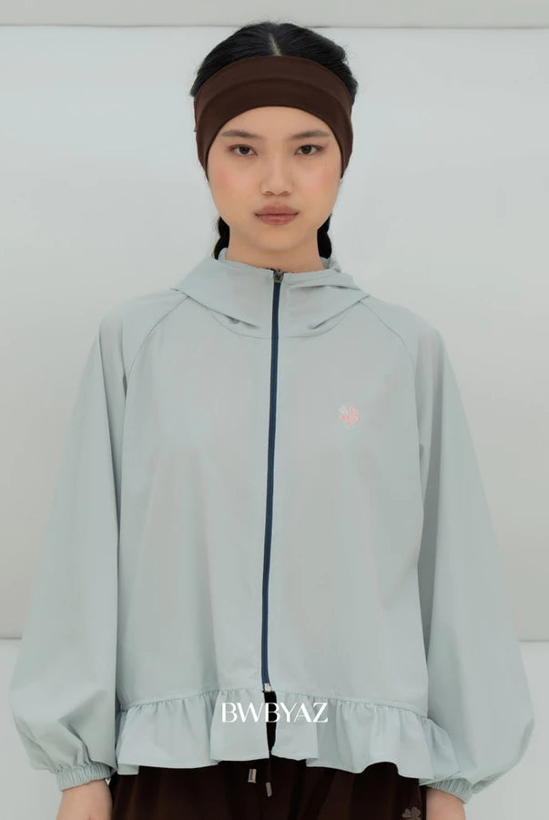 A model wearing a light awua blue hooded windbreaker jacket by BWBYAZ with a dark navy zipper, a small pink embroidered logo on the chest, and a ruffled hem. She is also wearing a dark brown headband and dark brown pants against a plain light gray studio background.
