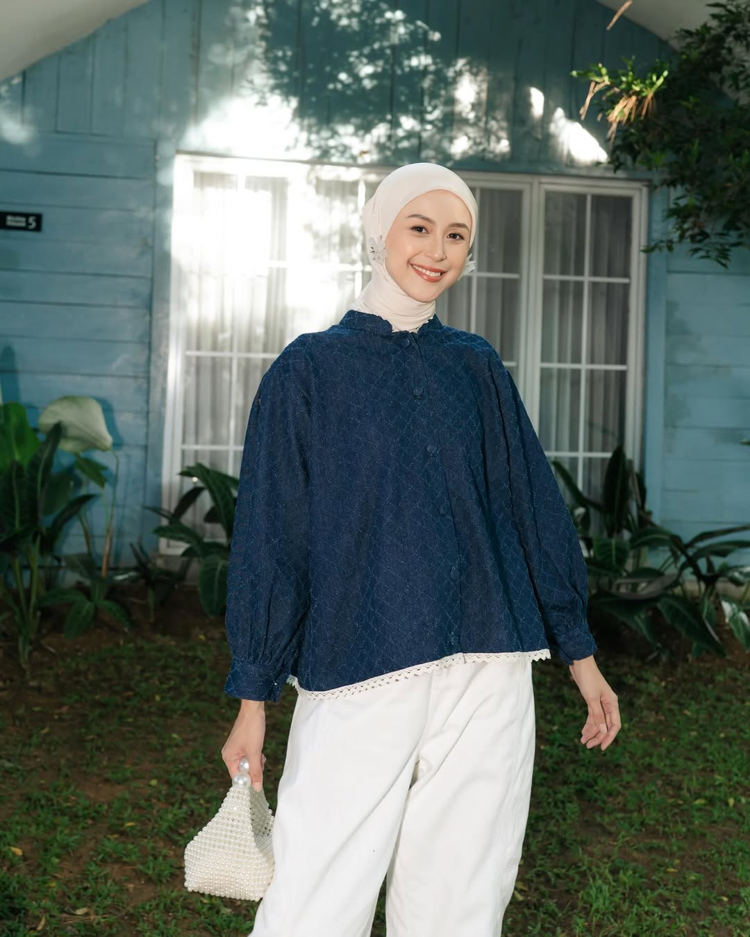 Woman wearing a navy blue top from Haidee Orlin and white pants standing in front of a light blue house with white windows.