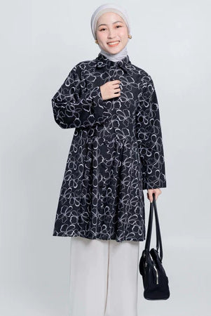 Woman wearing a black patterned tunic and white pants, holding a black bag on a white background