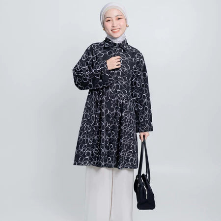 Woman wearing a black patterned tunic and white pants, holding a black bag on a white background