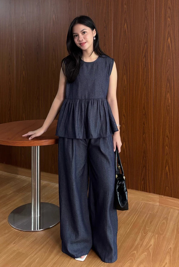 Woman in a dark denim top and pants by Renaette standing next to a wooden table against a wooden wall.