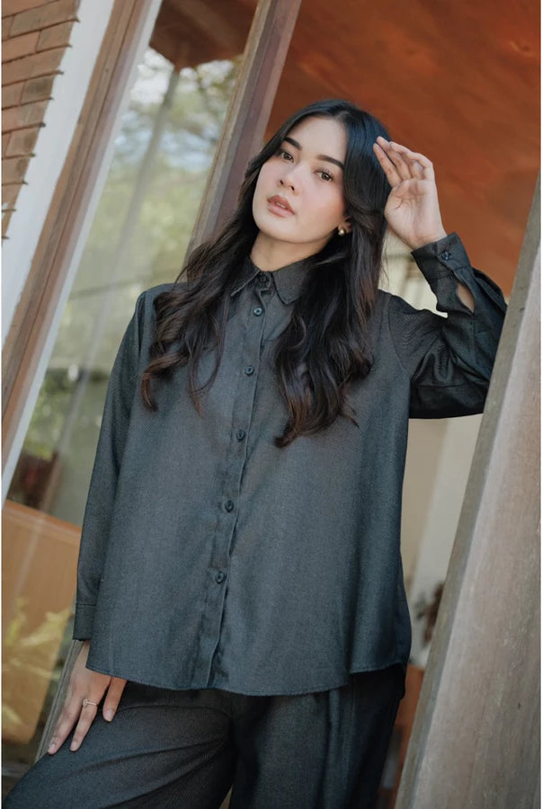 Woman wearing a dark grey shirt by Renaette and black pants standing indoors with a blurred background