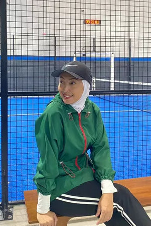 Person wearing a green track jacket by Abame and black cap sitting on a bench in an indoor sports facility.