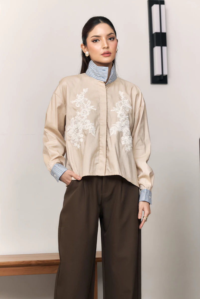 Woman wearing a beige shirt with floral embroidery by Studio Tui and brown pants against a plain background