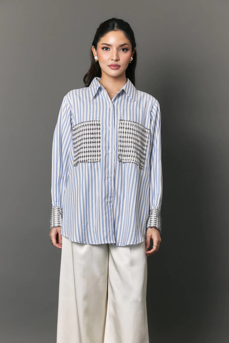 A woman modeling a light blue and white vertically striped button-down shirt by Studio Tui with decorative black-and-white patterned chest panels and striped cuffs, paired with wide-leg cream pants, against a dark gray studio background.