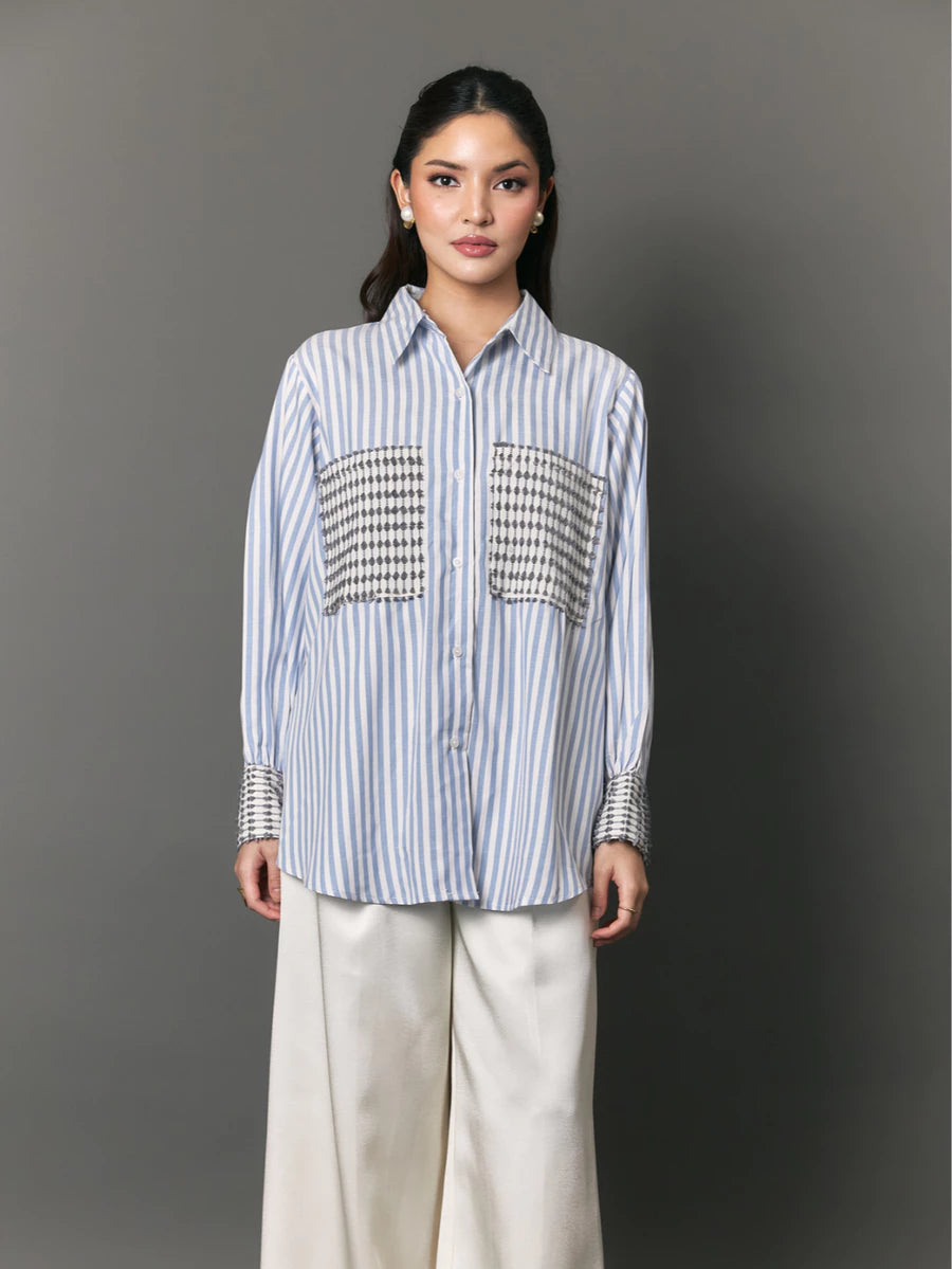 A woman modeling a light blue and white vertically striped button-down shirt by Studio Tui with decorative black-and-white patterned chest panels and striped cuffs, paired with wide-leg cream pants, against a dark gray studio background.