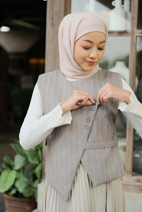 Woman wearing a gray pinstripe vest by Tunique over a white long-sleeve shirt, standing in an outdoor setting with plants.