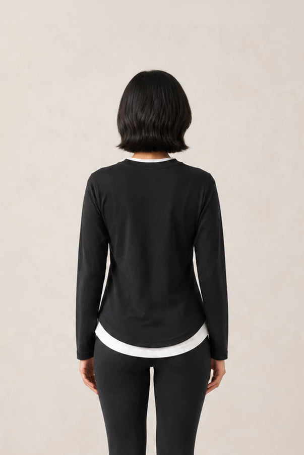 A woman viewed from behind wearing a black long-sleeve athleisure t-shirts by Jenna & Kaia with white contrast trim at the collar and hem, paired with black leggings, against a plain light beige background.