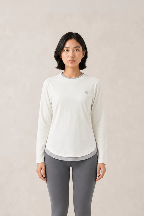 A woman modeling a white long-sleeve athletic top by Jenna & Kaia with a small embroidered logo on the left chest, paired with gray leggings, against a neutral beige textured background.
