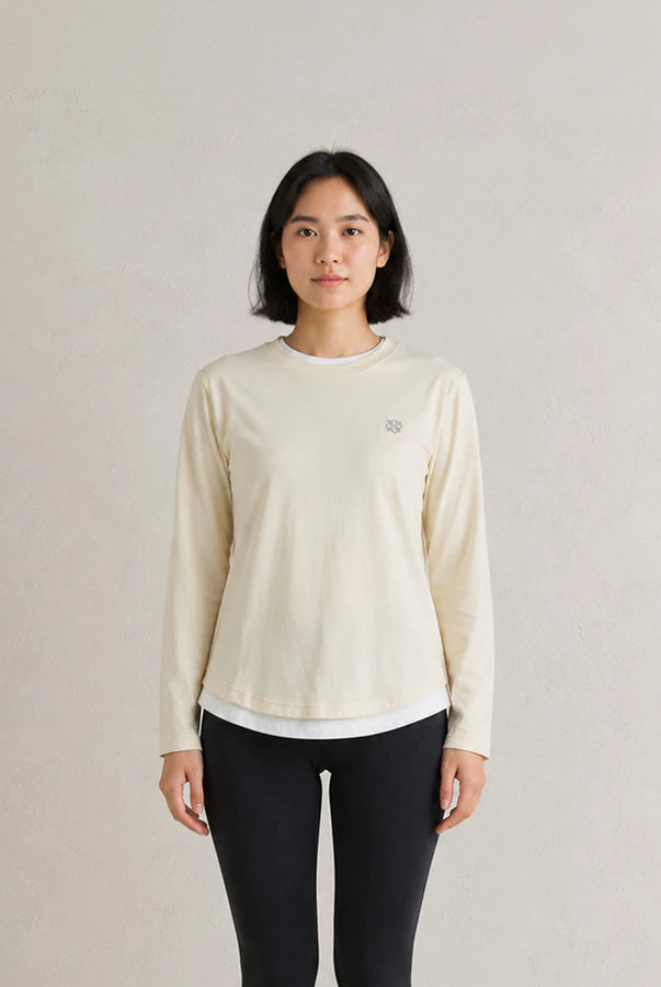 A woman stands against a light beige textured wall wearing a cream-colored long-sleeve athletic top by Jenna & Kaia with a small embroidered logo on the left chest, paired with black leggings.