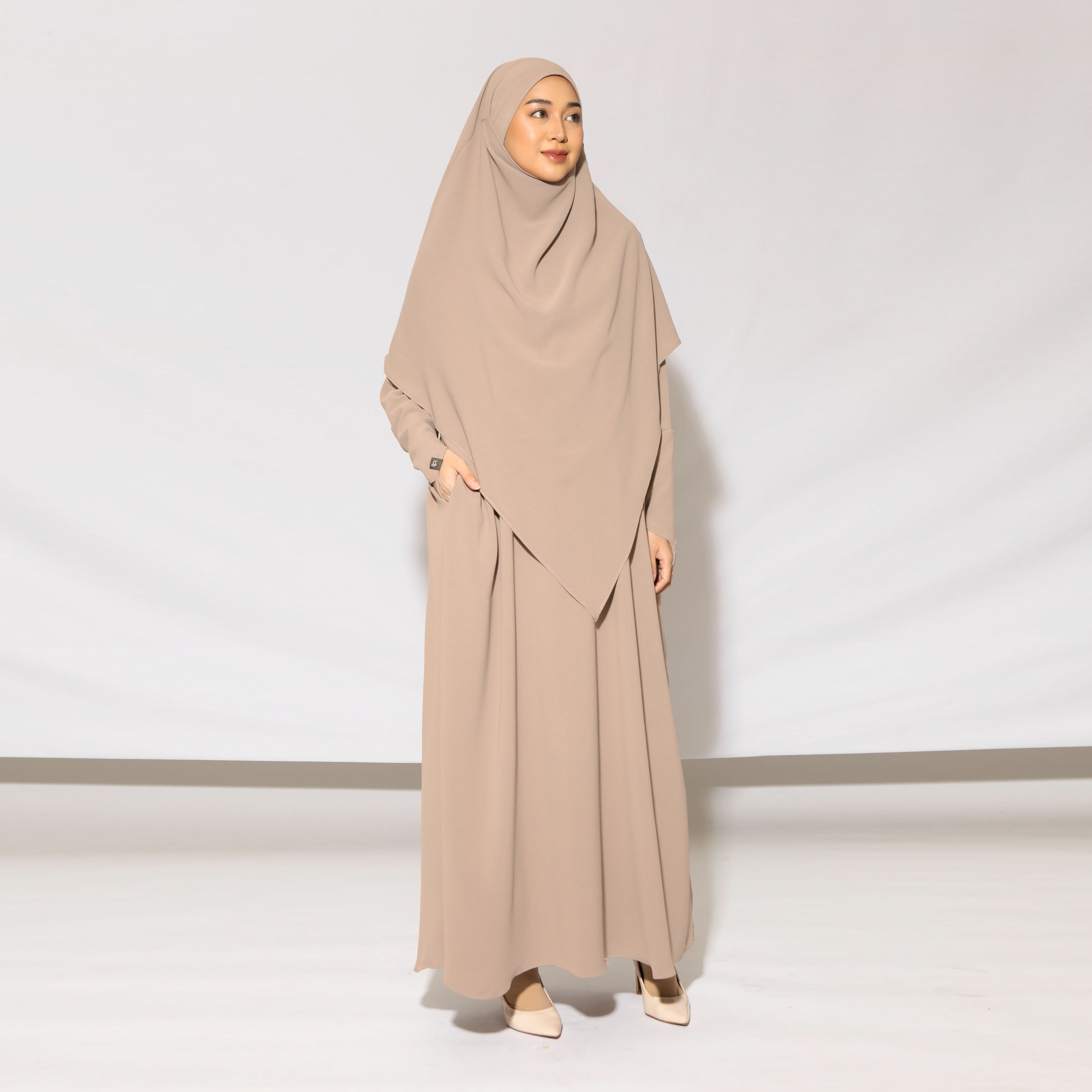 A woman standing in a studio wearing a beige khadija abaya set by Lozy Hijab with a matching headscarf, designed for coverage and elegance in a neutral color.