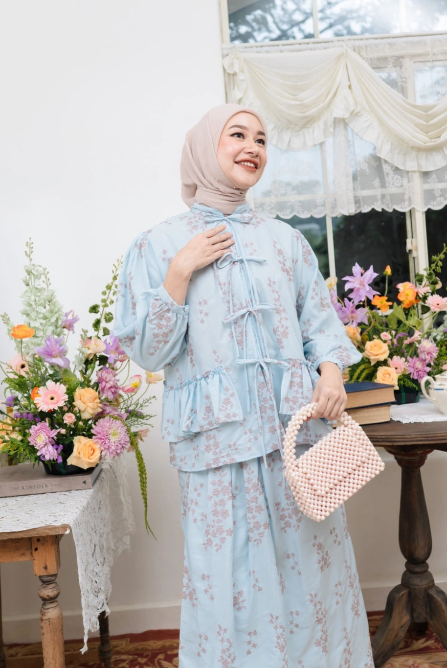 Woman in a light blue floral outfit by Haidee Orlin standing in a room with flowers and a table.