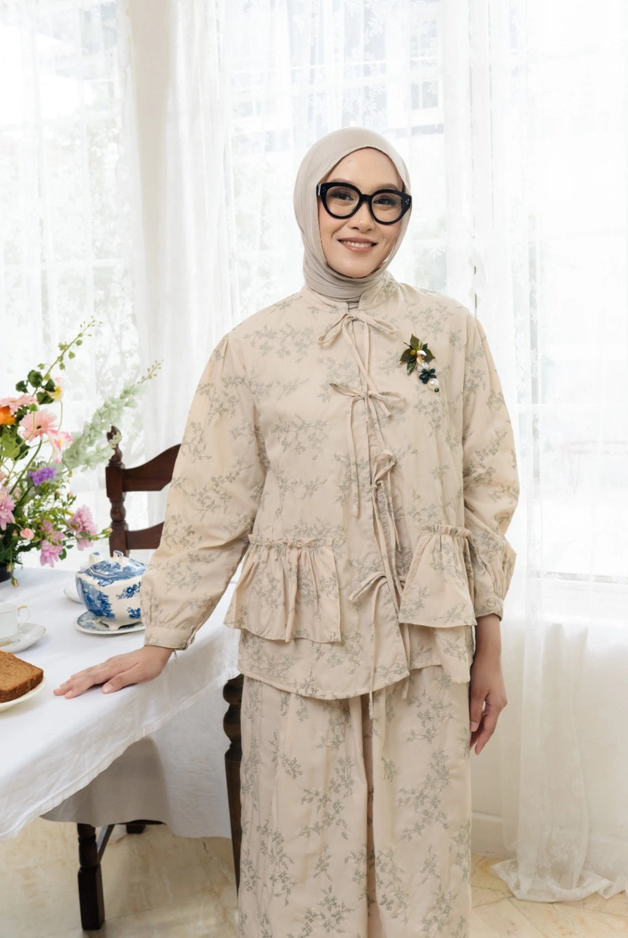 Person wearing a cream floral outfit by Haidee Orlin standing in a bright room with white curtains and a table with flowers.