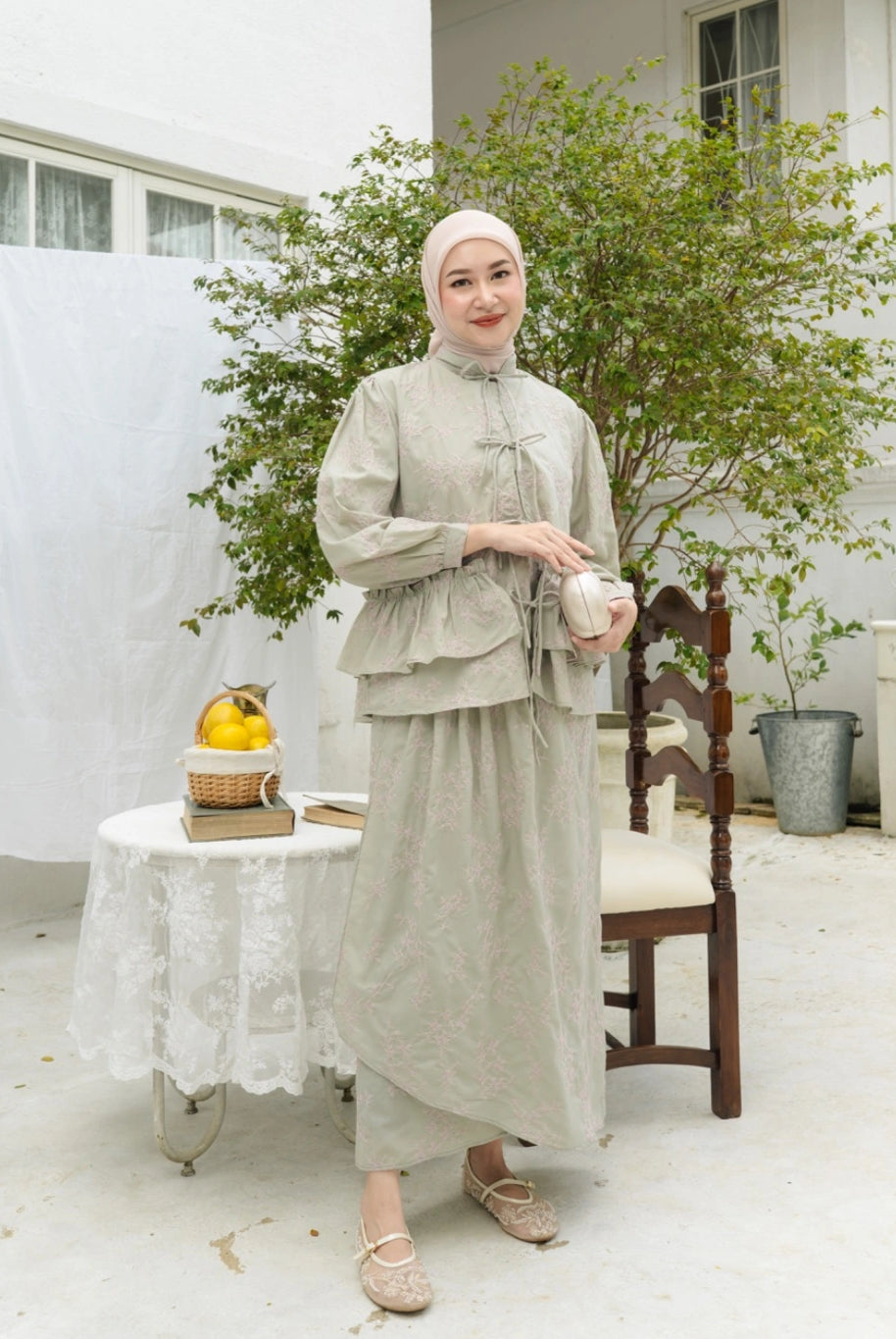 Woman in a light sage traditional outfit by Haidee Orlin standing in a bright room with plants and furniture.