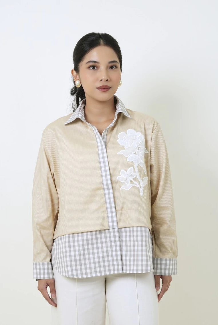 A woman modeling a beige jacket by Studio Tui with white floral embroidery on the chest and white and gray gingham layering, paired with white pants, against a plain off-white background.