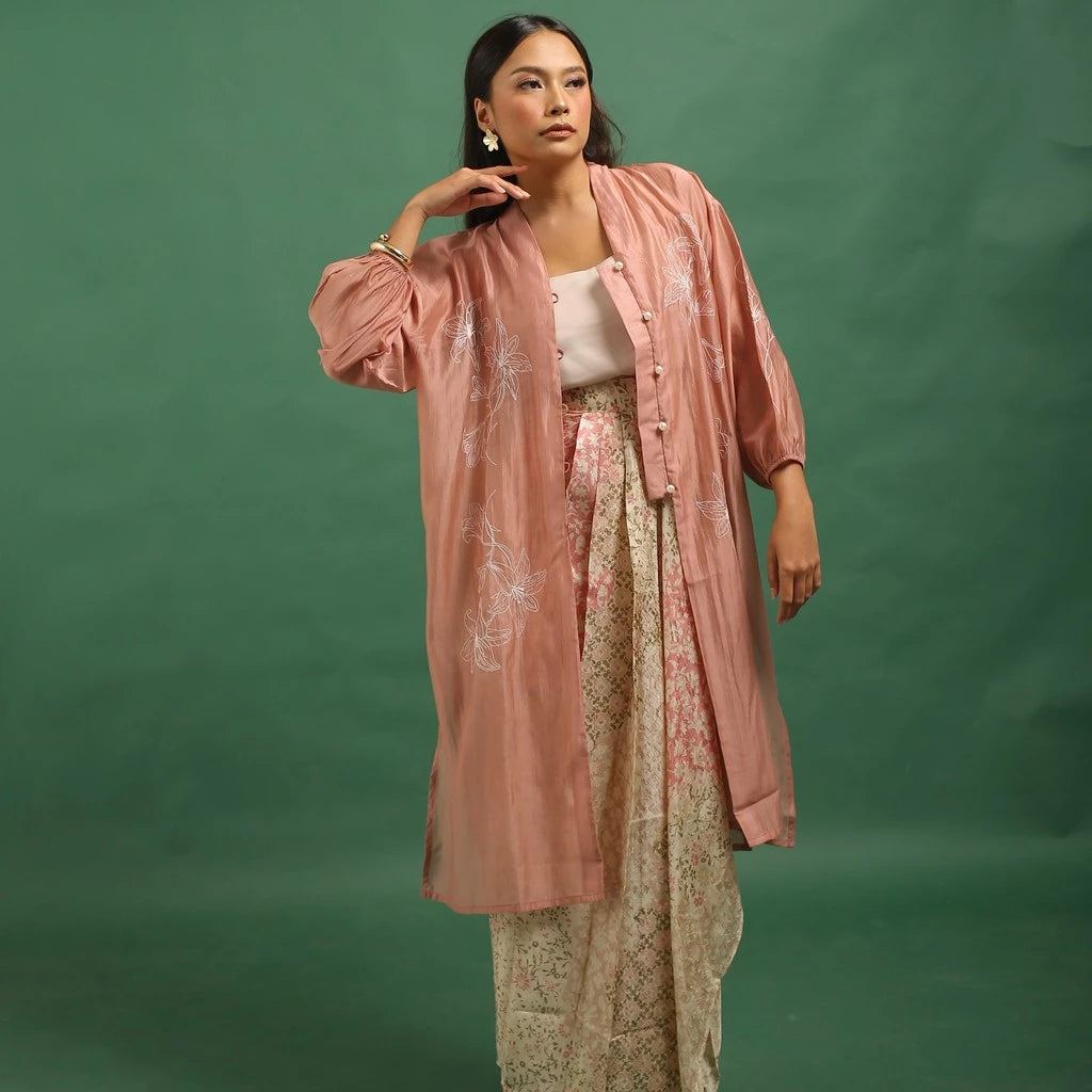 Woman wearing a coral colored embroidered kebaya by Kartinis Label and patterned pants against a green background