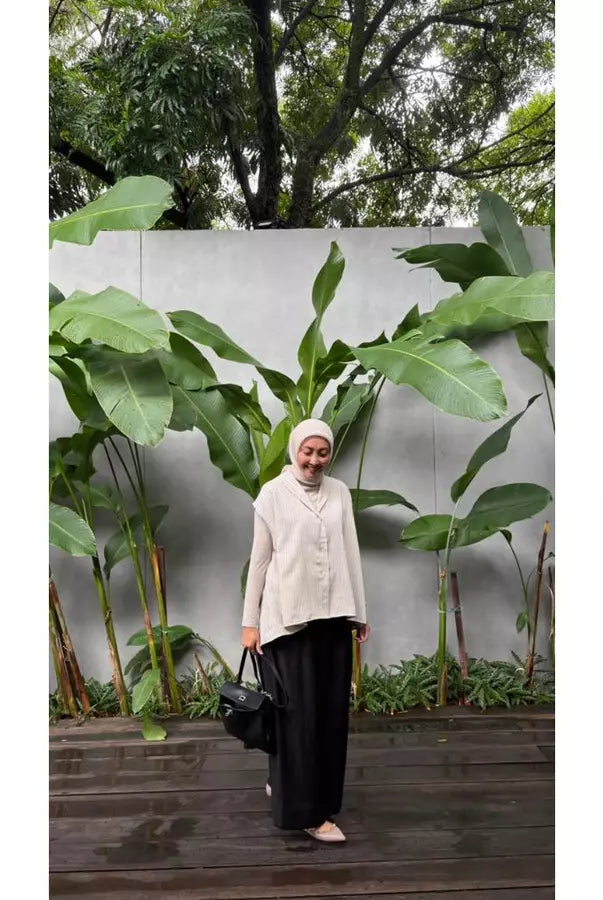 Woman in a beige vest by Abame over a beige shirt standing in front of a white wall with green plants