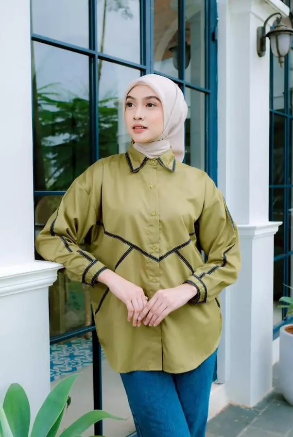 Woman wearing a green shirt by Abame and blue pants standing in front of a white building with glass doors.