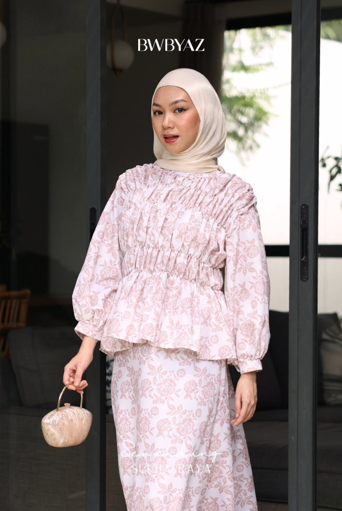 A woman wearing a pink floral blouse with smocked details on the chest and waist from BWBYAZ, and a matching pink headscarf. She is holding a small purse and standing in front of a glass door.
