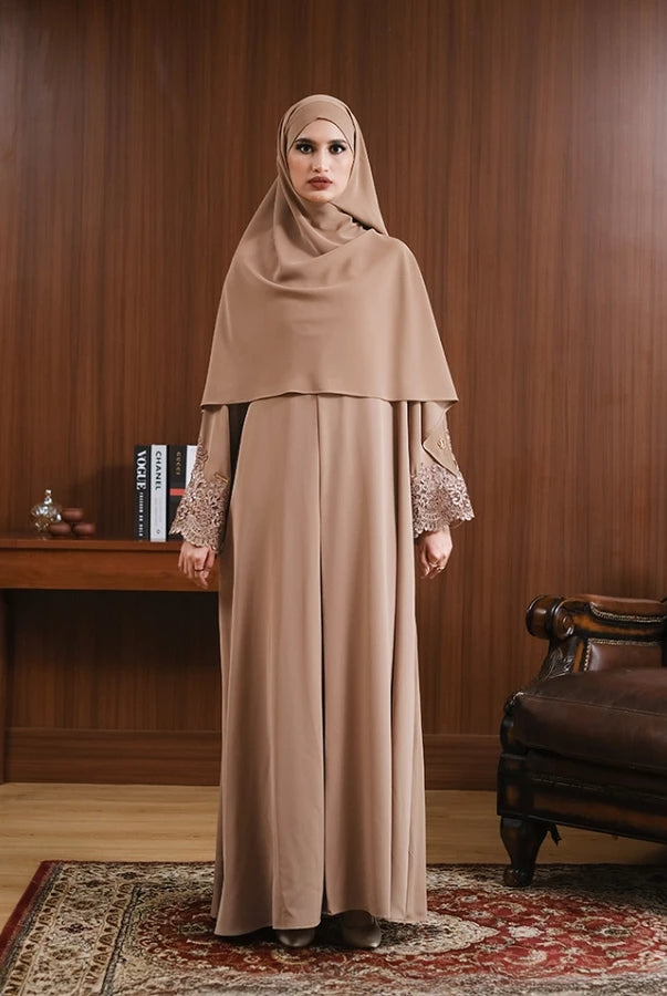 Woman in a brown hijab and abaya by Tubita standing in a room with wooden walls and a leather chair.