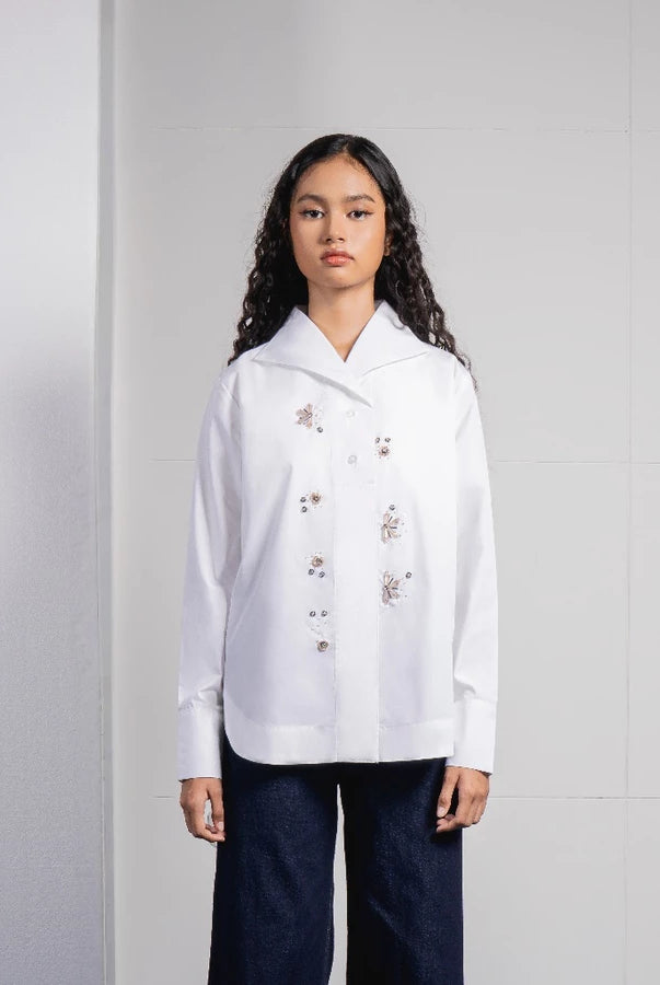 A model stands against a light gray tiled wall wearing a white long-sleeve button-up shirt by Jenna & Kaia with delicate floral embroidery on the front and dark navy wide-leg pants.