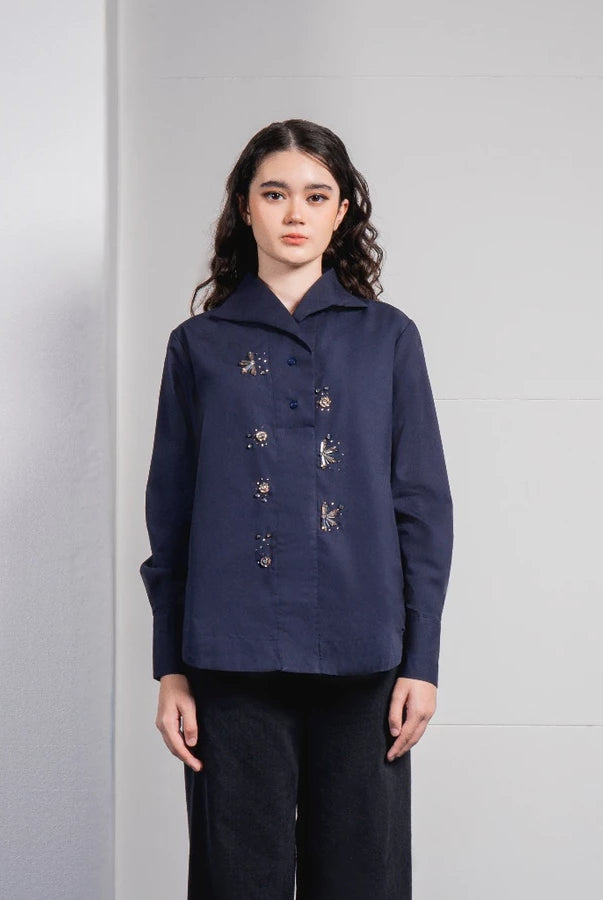 A woman stands against a light gray tiled wall wearing a dark navy long-sleeve button-front shirt by Jenna & Kaia with small embroidered details and dark wide-leg pants.