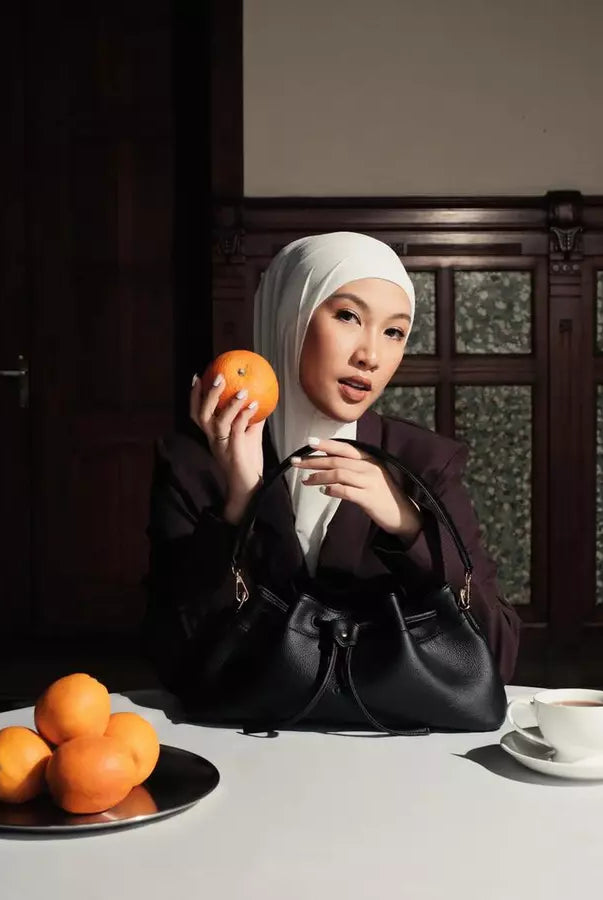 Woman holding black shoulder bag by Prior at a table with oranges and a cup of coffee.