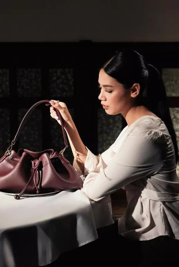 Woman sitting at a table with a burgundy shoulder bag by Prior in a dimly lit room