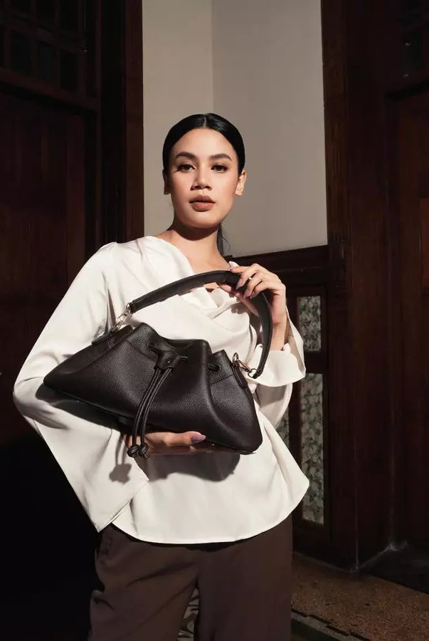 Woman holding a dark brown shoulder bag by Prior in a dimly lit room